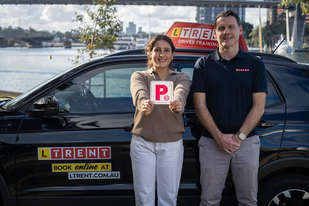 Learner driver with P plate standing beside an LTrent driving instructor and training car.
