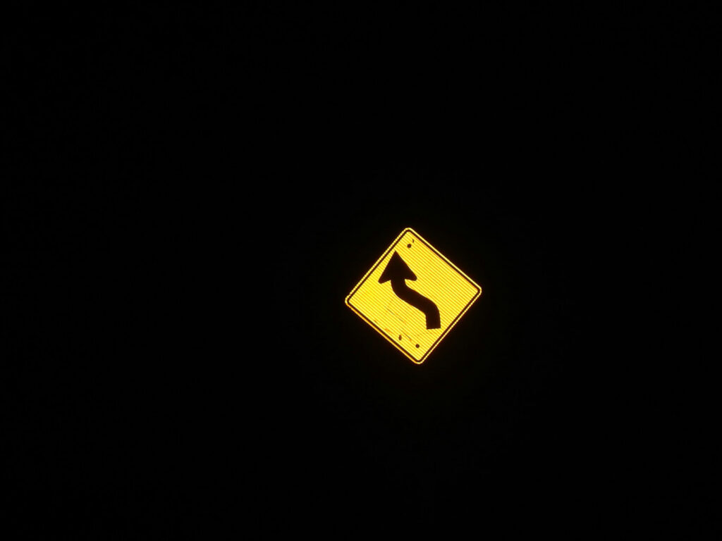 Reflective yellow road sign showing a winding road symbol against a dark background.
