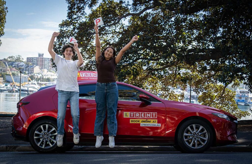Two learners holding red P plates and celebrating beside an LTrent driving school car.
